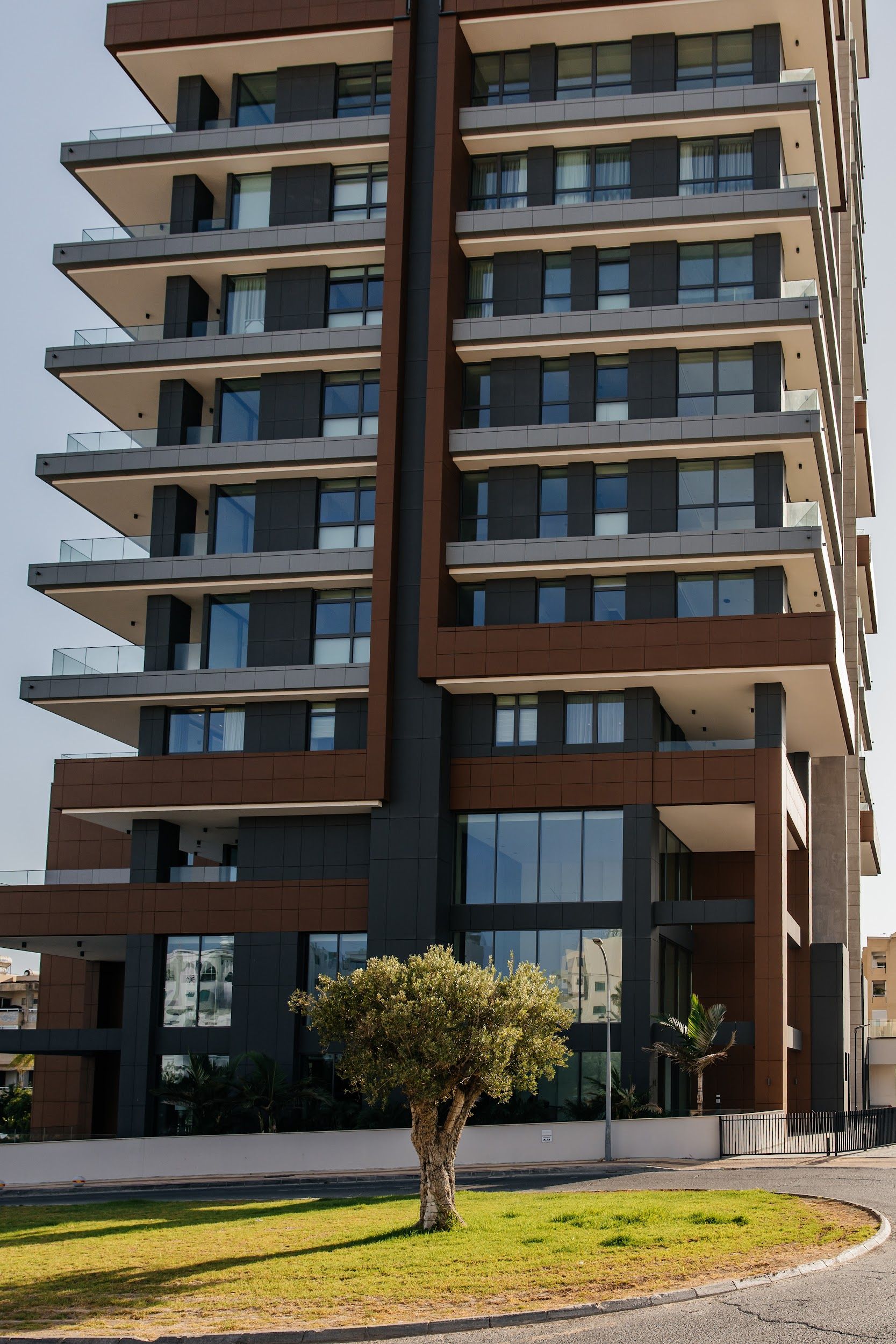Modern high-rise apartment building with large balconies, a small tree, and green grass.