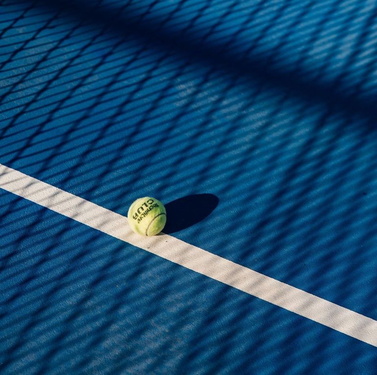 Yellow tennis ball on blue court, resting on a white line with net shadows.
