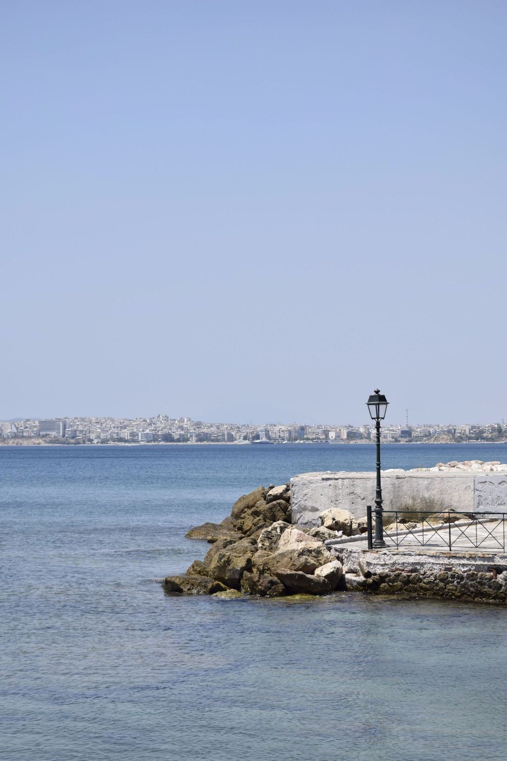 A stone pier with a lamppost extending into blue water under a clear sky.