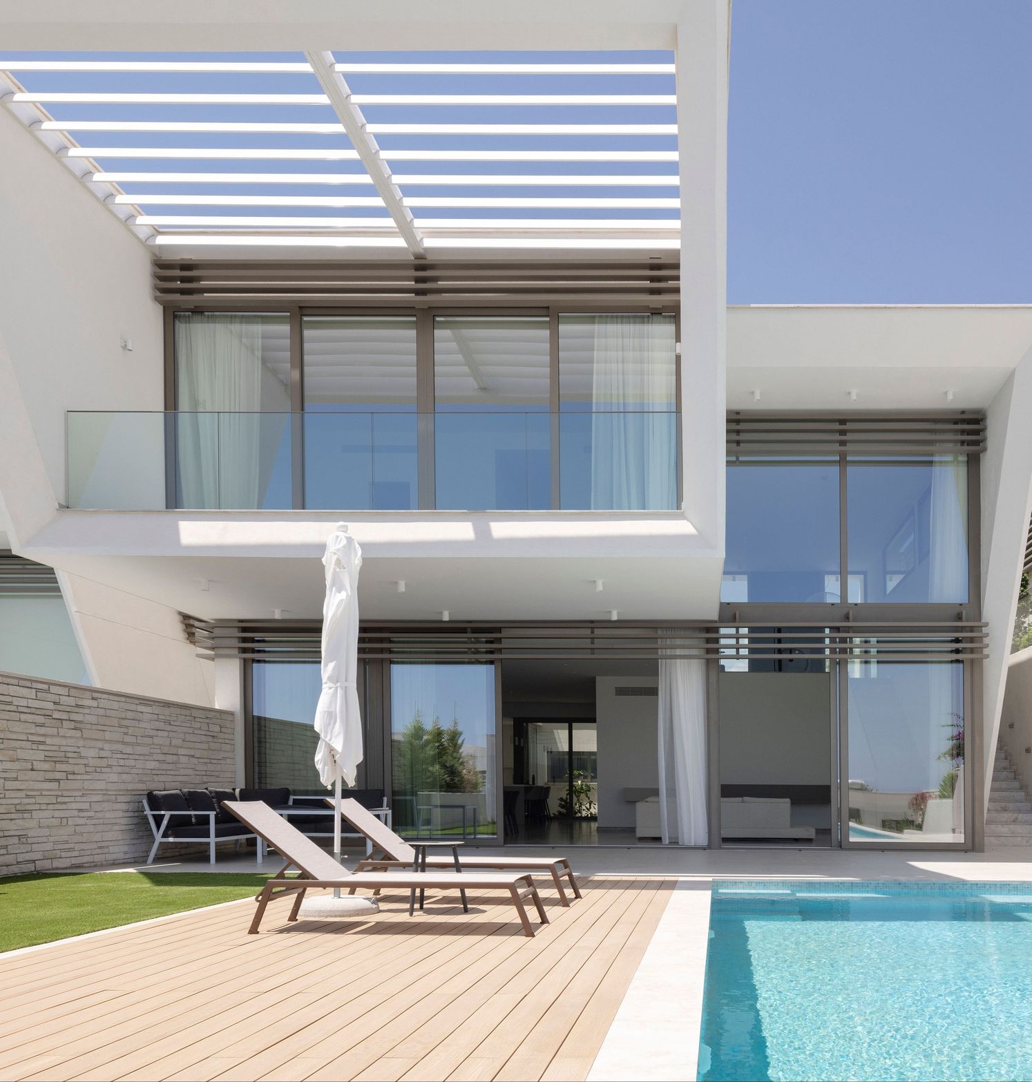 Modern white house with large windows, swimming pool, wooden deck, and lounge chairs.