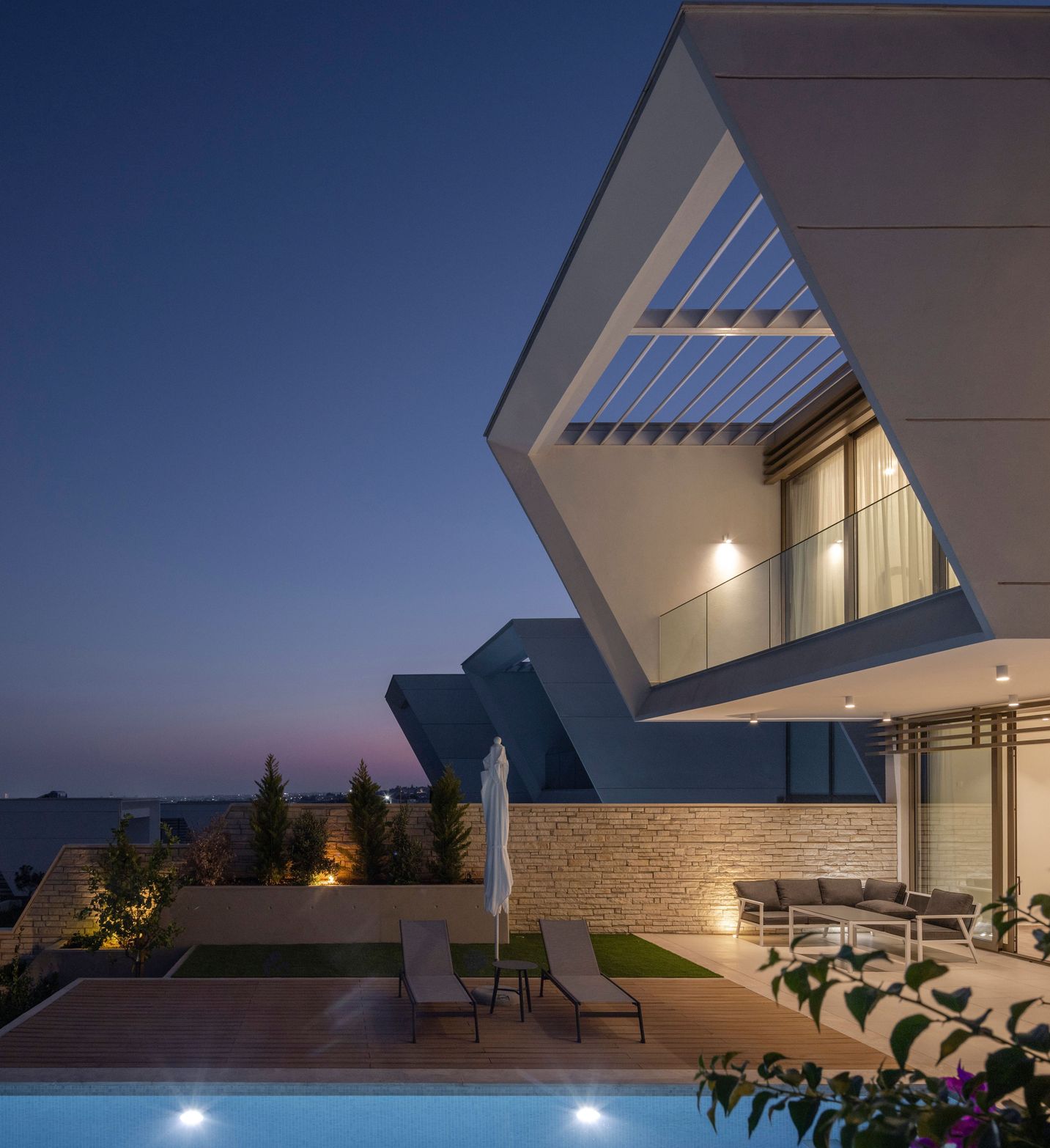 Modern geometric white house with a lit swimming pool and patio at twilight.