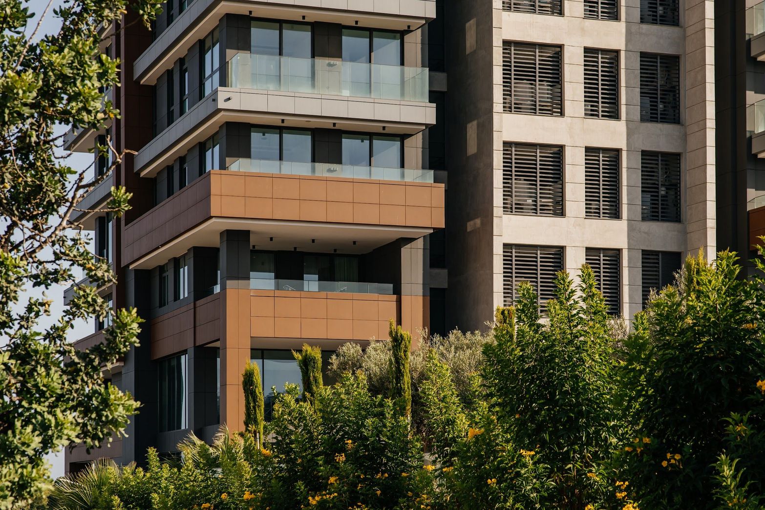 Modern apartment building with glass balconies and wood-paneled sections amidst lush green trees.