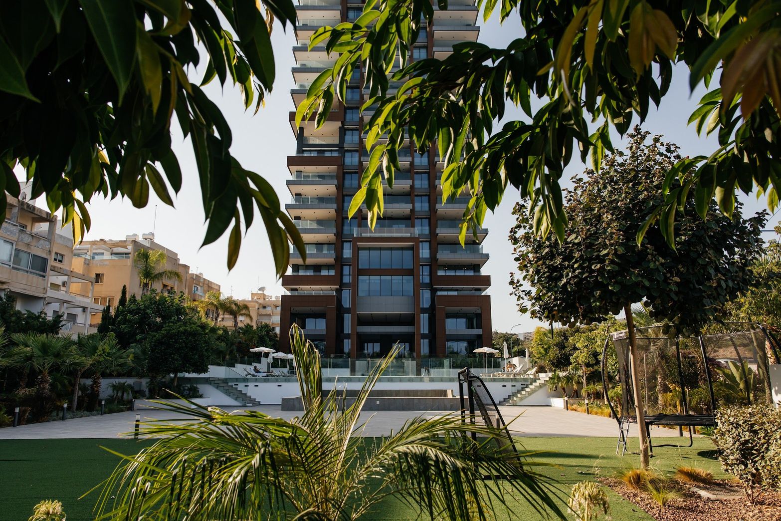 Modern high-rise building with many balconies, framed by lush green foliage and a manicured lawn.