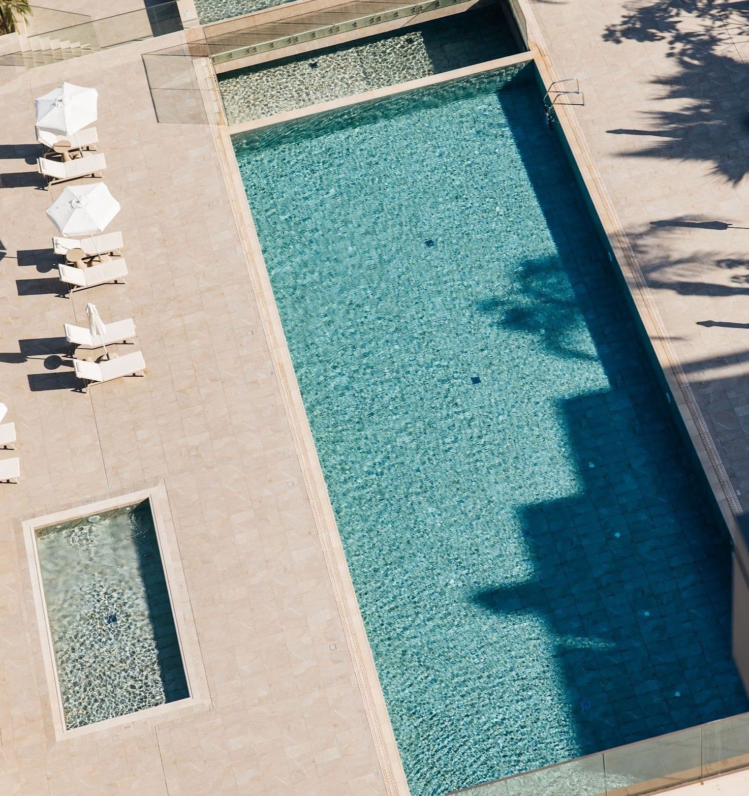 Aerial view of a rectangular turquoise swimming pool with white lounge chairs on light paving.