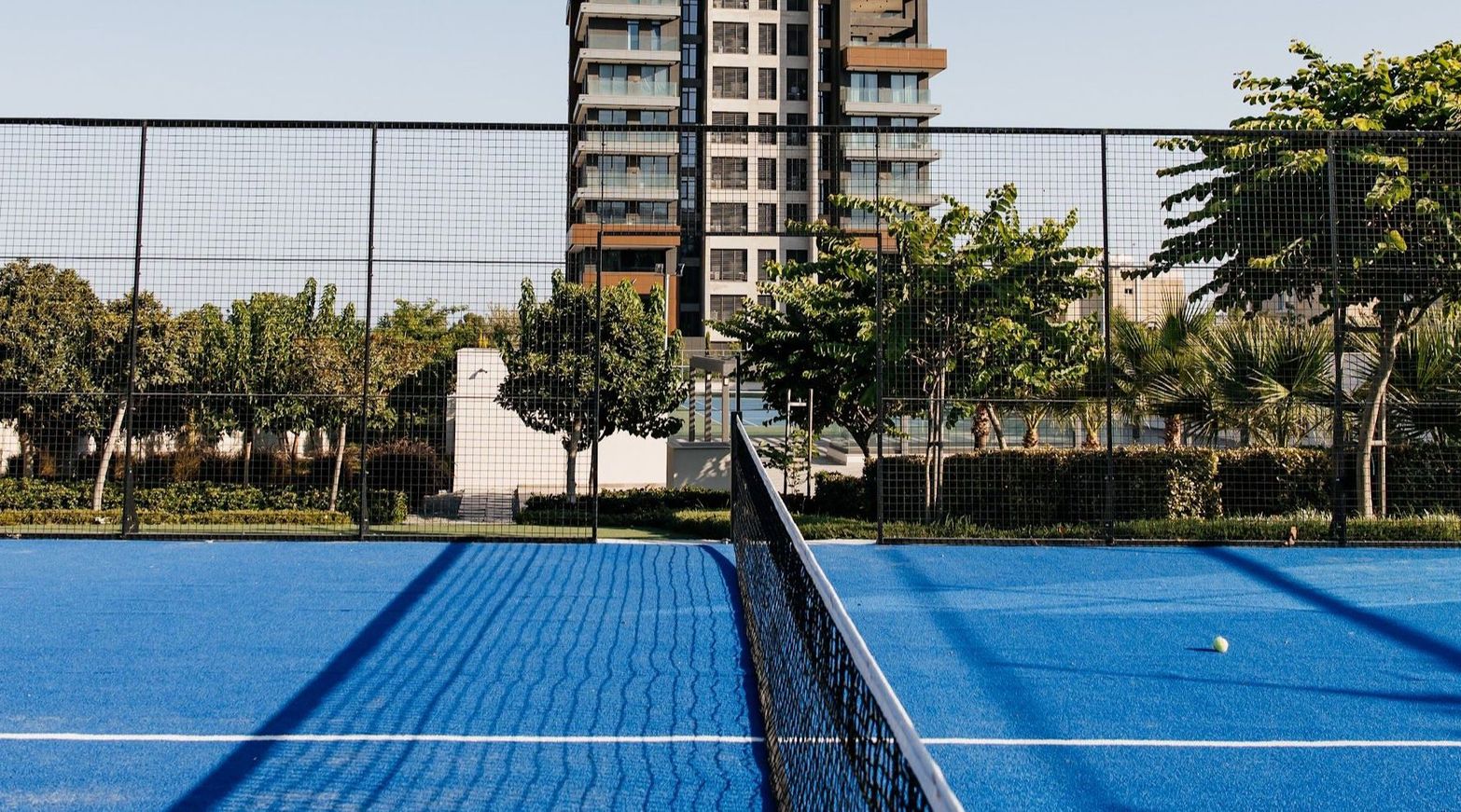 Blue tennis court with a net and white ball in front of a modern building and trees.