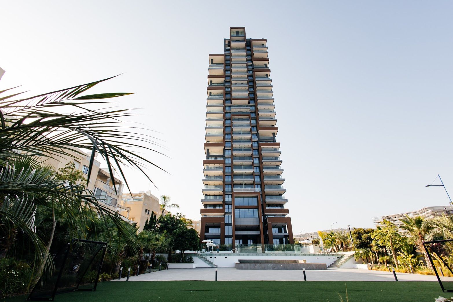 Modern high-rise building with numerous balconies, surrounded by green landscape under a clear sky.