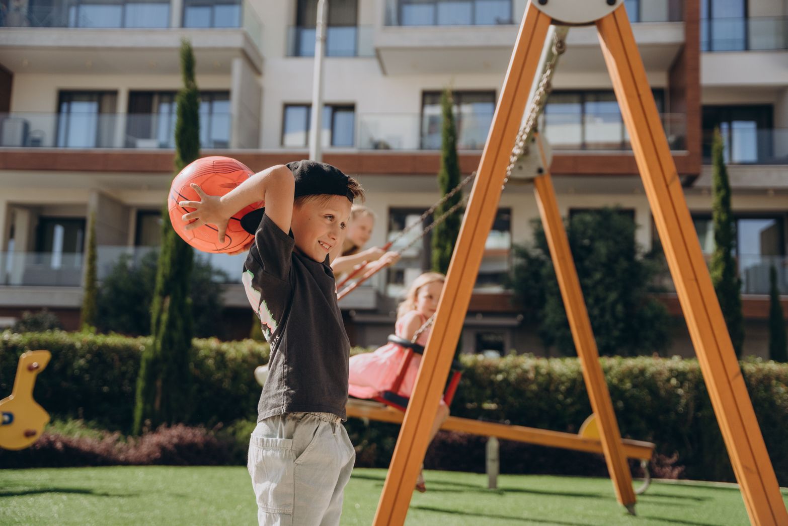 Young boy plays with an orange ball on a sunny playground near a swing set.