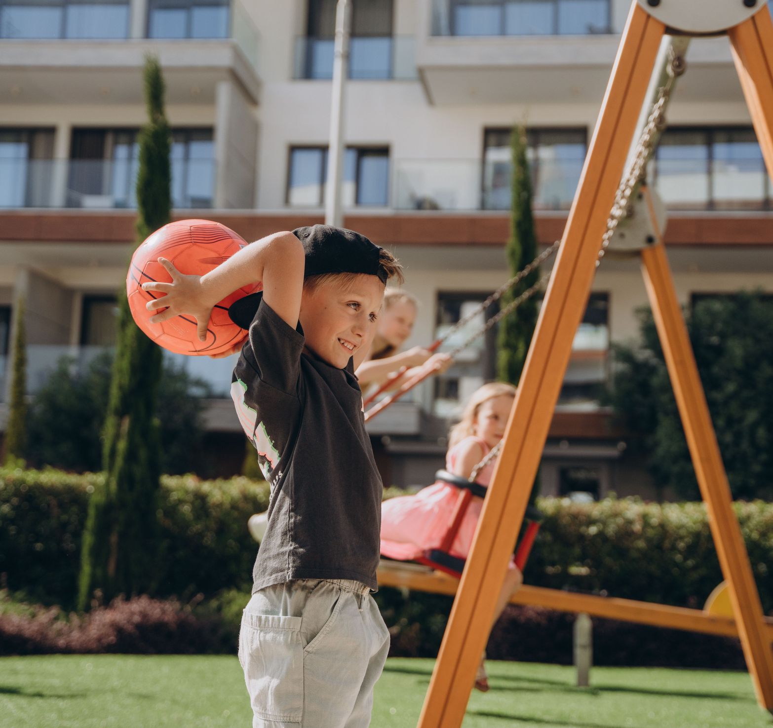 Smiling boy holds orange ball, girl on swing, children playing happily outdoors.
