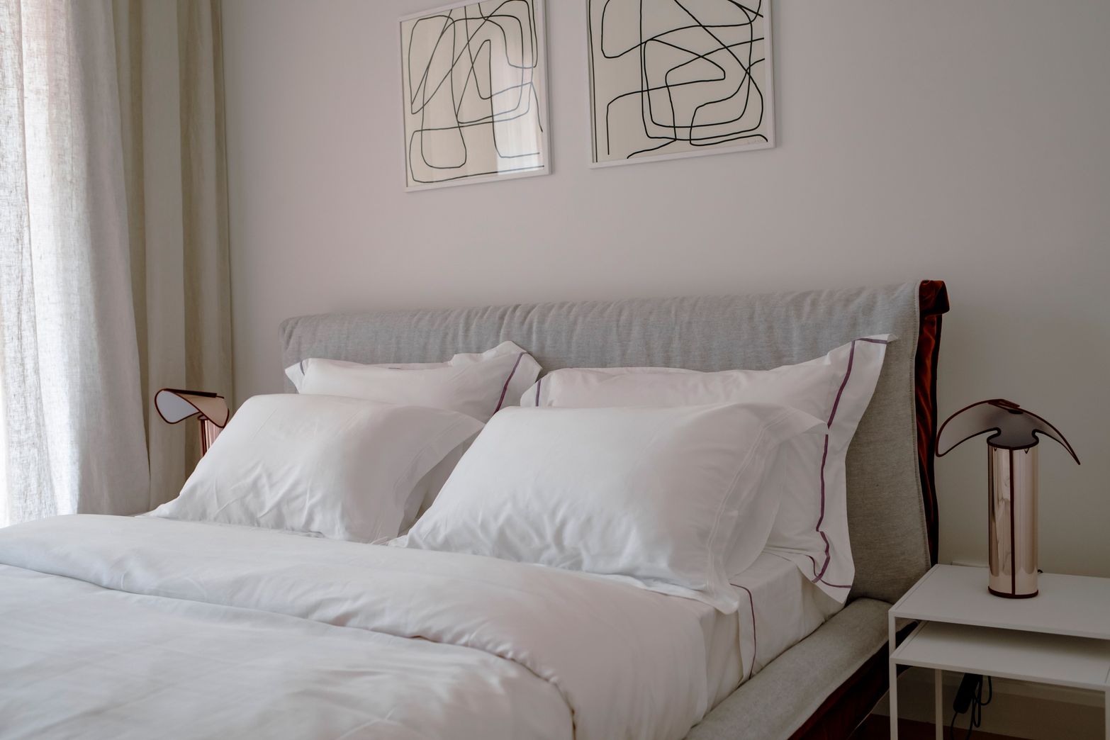 Modern bedroom with a bed, white bedding, a light grey headboard, and minimalist abstract wall art.