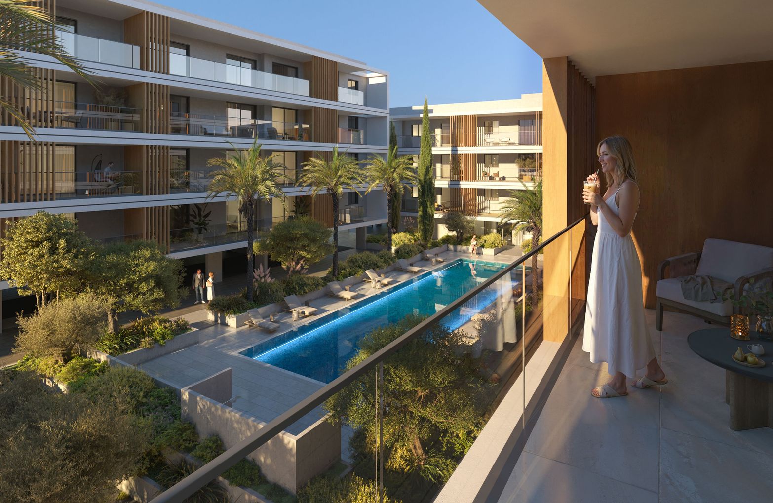 Woman on balcony overlooking a modern resort with a long swimming pool and lush gardens.