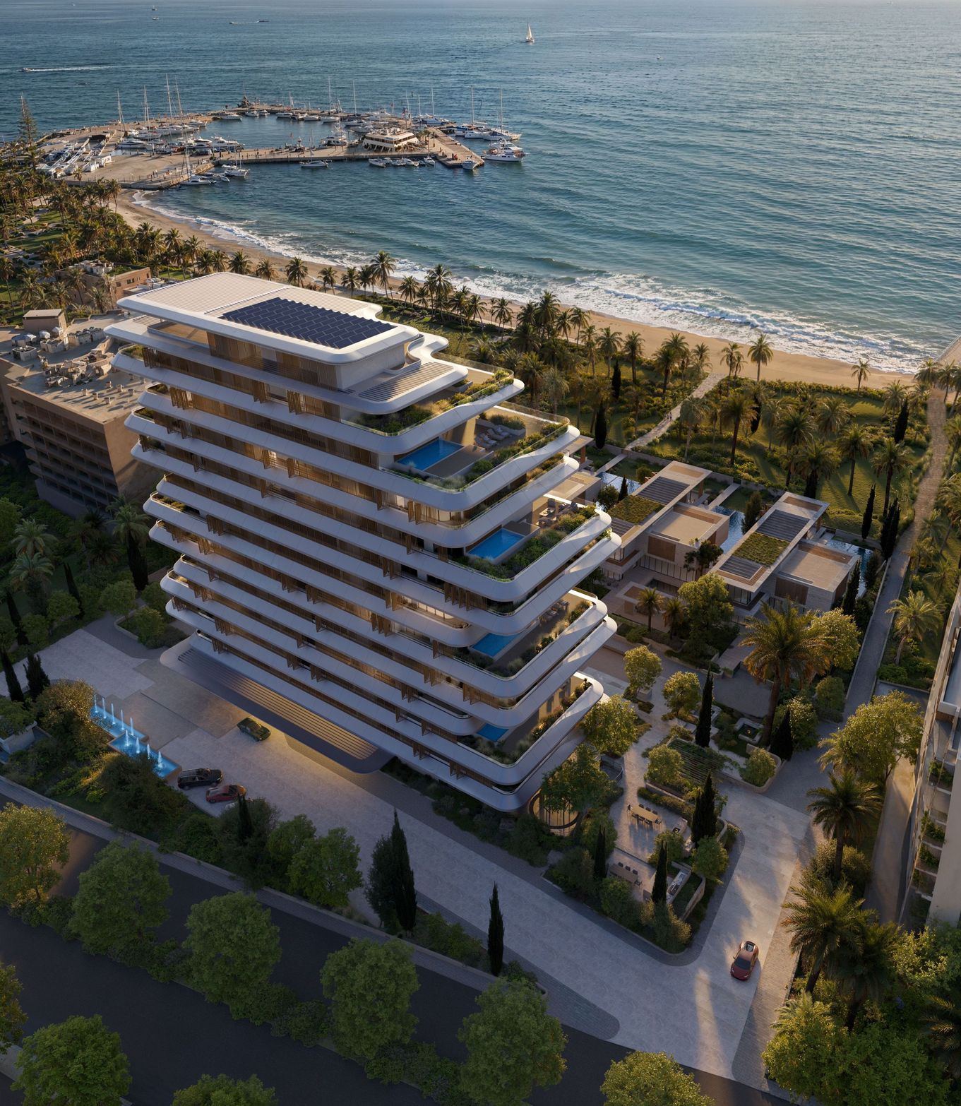 Modern, stacked beachfront building with green terraces overlooks a marina and sandy coastline.
