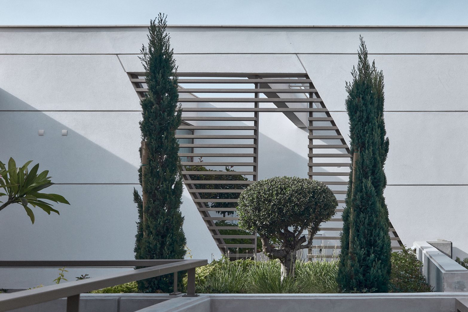Modern white building facade with geometric design and tall cypress trees in foreground.