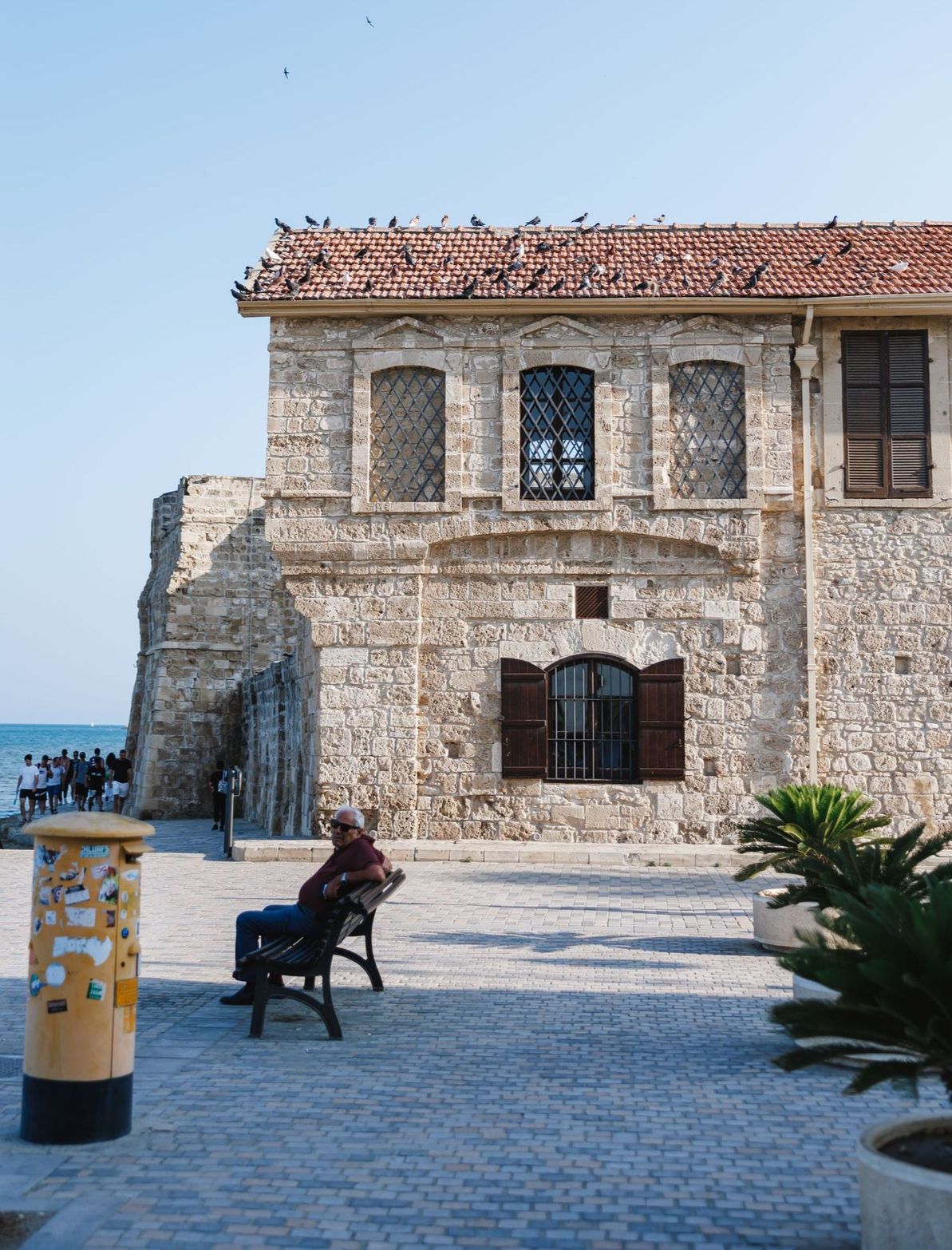 Historic stone building with a red tiled roof, boarded windows, and a person on a bench by the sea.