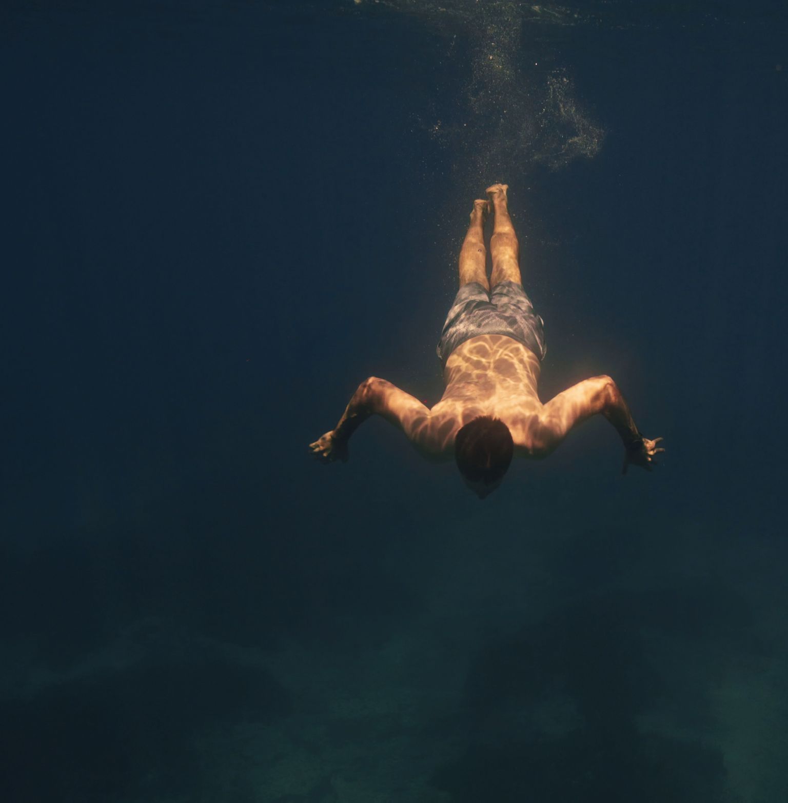Man swimming underwater in dark blue water, seen from below with bubbles.