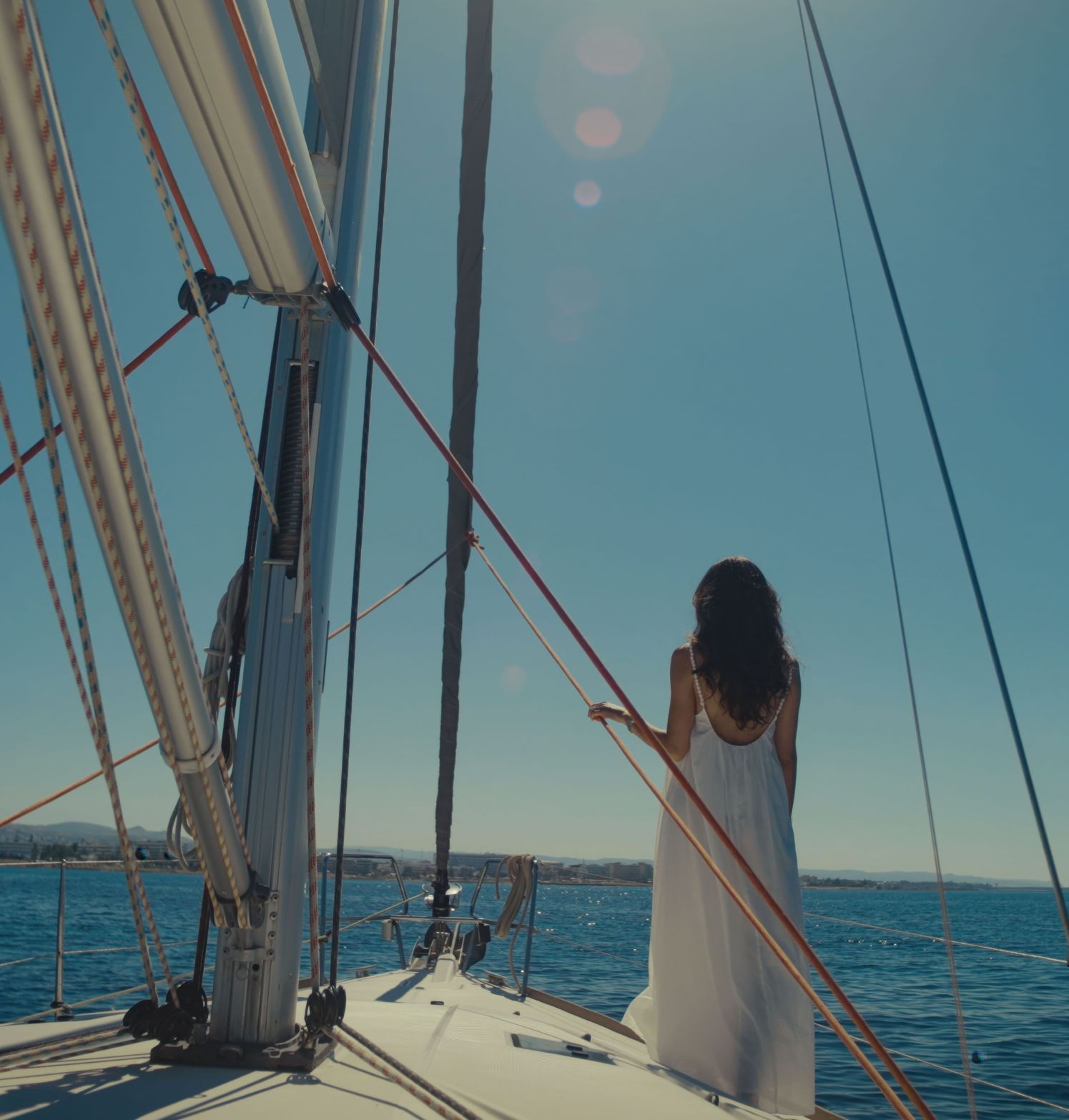 Woman in white dress on sailboat gazes at bright blue ocean under sunny sky.
