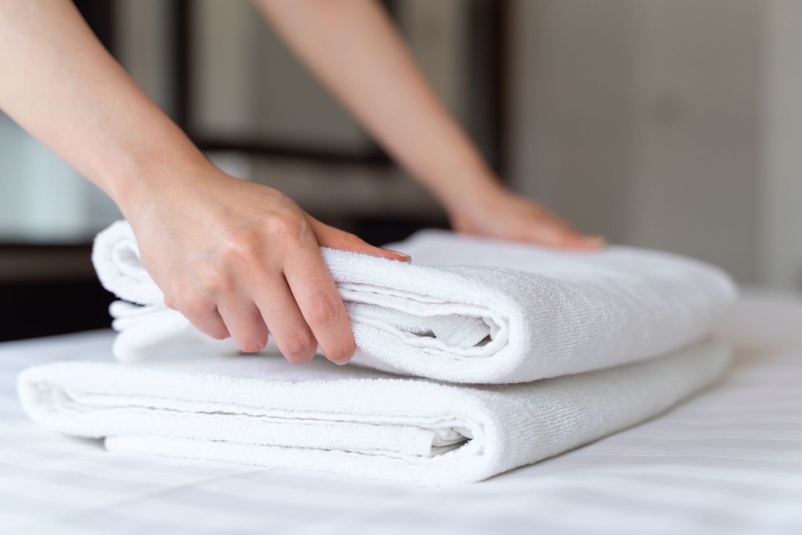 Hands neatly folding clean white towels on a bed, demonstrating housekeeping. 